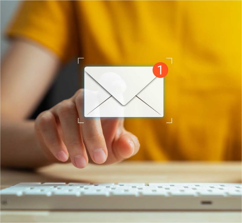 Person in a yellow t-shirt pointing at a mail envelope graphic with a notification, sitting in front of a keyboard.